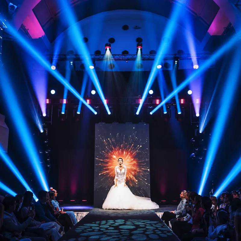 A model in a white wedding gown on a runway beneath blue stage beams, with the audience seated on both sides.