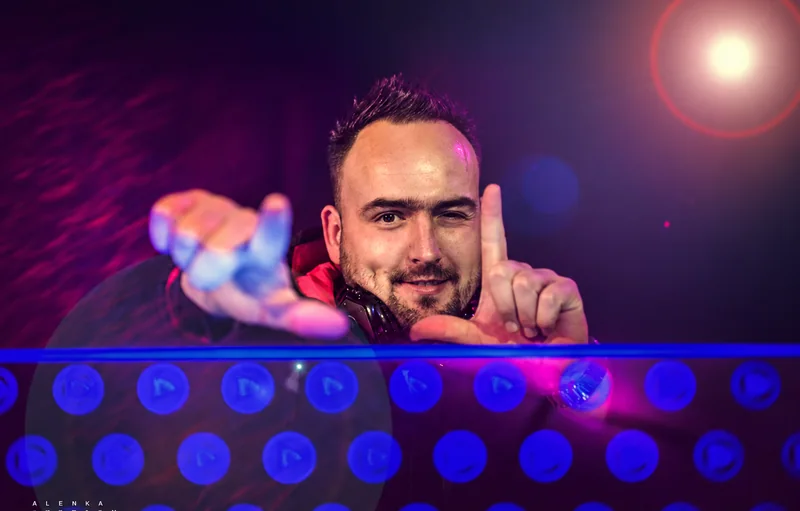 Tomy winking and gesturing toward the camera behind a DJ booth under purple and pink party lights.