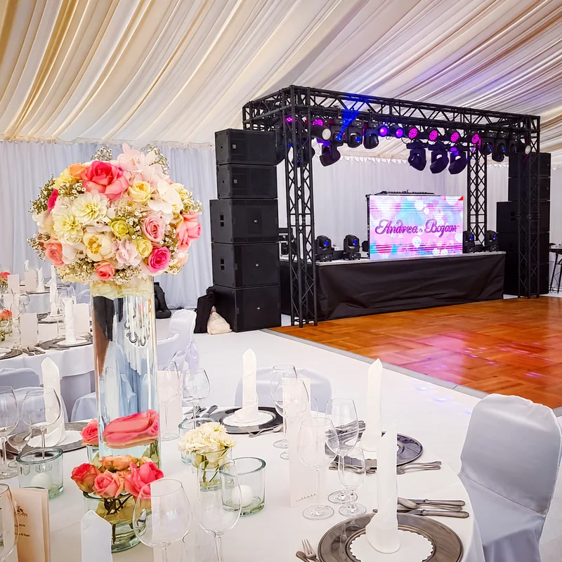 A wedding table with pastel flowers and glassware beside an empty dance floor, with a DJ stage and lighting rig inside a white marquee.