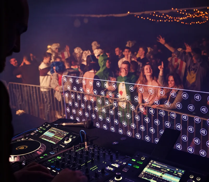 A view across DJ decks and a mixer toward a cheering crowd, with warm strings of lights overhead.