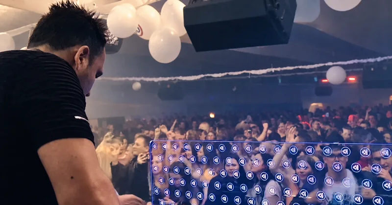 Video cover: a view from behind Tomy at the DJ booth toward a packed crowd, with hanging spheres above the dance floor.