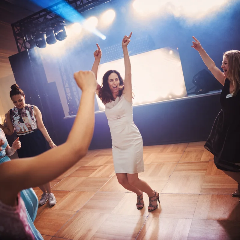 A woman in a white dress dancing with both arms raised, surrounded by other guests and bright stage lights.