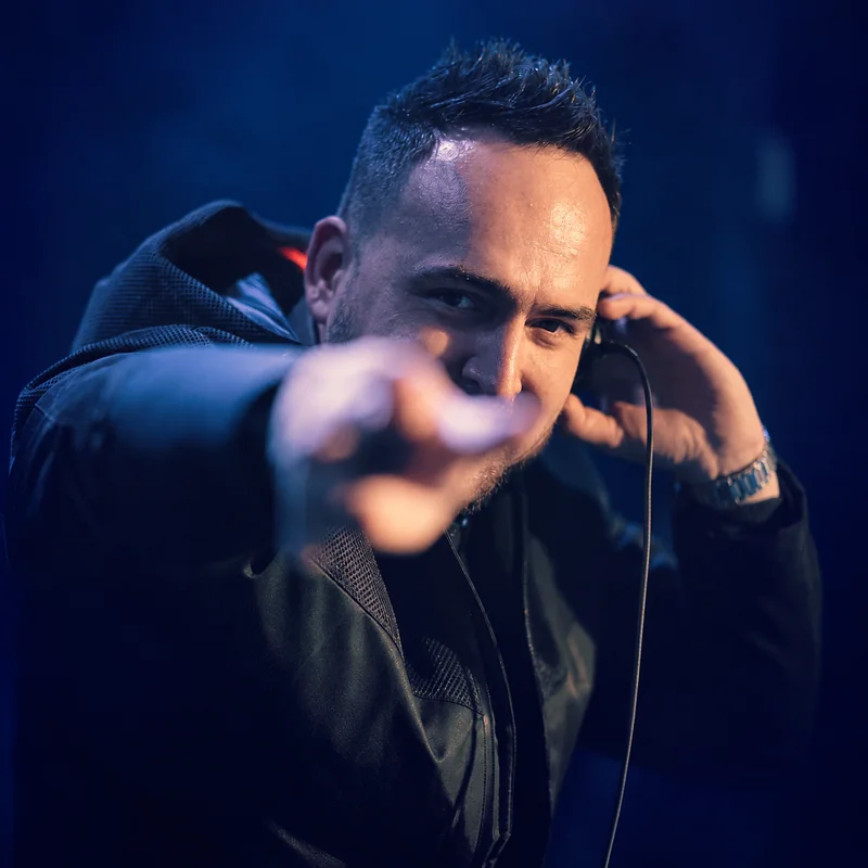 Tomy holding headphones to one ear and pointing toward the camera under blue DJ lighting.