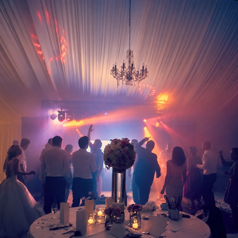 Wedding guests dancing beneath a chandelier and draped marquee ceiling, with flowers in the foreground and colourful lights behind them.