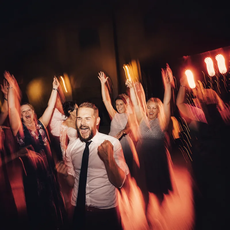 Wedding guests cheering with raised arms, captured with flowing warm light trails on the dance floor.