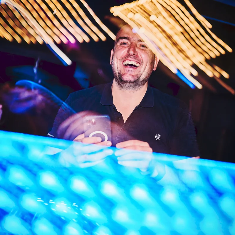 Tomy smiling behind a turquoise-lit DJ booth, framed by golden light trails.